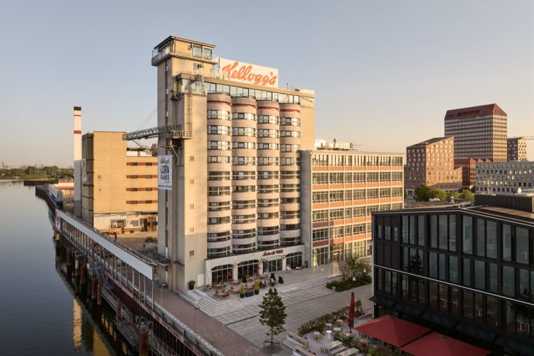 From grain silo to striking hotel building. For around four decades, the silo building in Bremen's Überseestadt served as a storage and production facility for the American Kellogg's corporation's cereal production. The towers, which date from the 1970s and are up to 40 metres high, stored several thousand tonnes of corn, rice, wheat and oats. The building was functional, solid and designed exclusively for industrial use. When operations ceased in 2017, the iconic building faced a fundamental question: how could such a monumental building be meaningfully integrated into the future? The answer lies in the consistent conversion and further development of the existing structure. In the course of the transformation of the former port and industrial areas into today's Überseeinsel, the silo building was not demolished, but deliberately preserved and reinterpreted. A closed production site was turned into an open, usable space for people. Today, the former silo tubes house an extraordinary hotel. The use of the building is complemented by office, conference and restaurant space, as well as other publicly accessible facilities. As a striking high point, the Kellogg's Hotel now forms the core of the new city centre on Überseeinsel and plays a central role as a place of encounter and orientation. The new function brings the industrial origins of the building to life and at the same time gives it a completely new meaning in the everyday life of the city. Instead of empty industrial buildings, a lively place has been created. Kellogg Pier contributes significantly to the revitalisation of Überseeinsel and makes industrial history accessible to a wide audience. As part of the conversion, the former silo has also been modernised to improve its energy efficiency. Insulating the round silo shapes presented a particular technical challenge. In particular, the necessary compensation for the strongly curved surfaces required the utmost precision in the execution of the composite thermal insulation system. These complex geometries were implemented in a technically safe and creatively sophisticated manner. A creative plastering technique gives the façade a dynamic, mineral look that both reflects its industrial character and conveys a modern impression.

Baumit insulation system
 Baumit Mineral

Baumit Materials
 ProContact / ProContact DC 56 / KlebeSpachtel
 Multi MC 55 W / MC 55 W / MultiContact MC 55 W
 Multi HM 50 / HaftMörtel HM 50
 MultiFill LTM 81
 PremiumPrimer / PremiumPrimer DG 27
 SchraubDübel S (STR)
 PuraColor / PuracrylColor
 MineralTherm 035 Light plus
